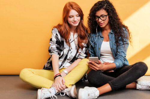 Two young women listening to a phone together - Best Podcasts Featured Image